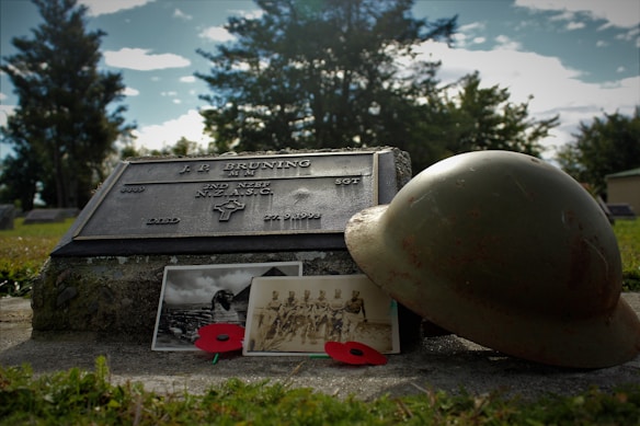 A gravestone with an inscription for a military individual is accompanied by a vintage helmet, black and white photographs, and two red poppies. Trees and a cloudy sky form the background, creating a serene and reflective setting.
