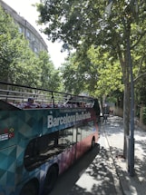 A colorful, double-decker tour bus labeled 'Barcelona Bus Turistic' is parked on a tree-lined street. The bus features an open top with vibrant seating visible. Tall trees cast shadows onto the sidewalk and street, while buildings with classical architecture can be seen in the background.