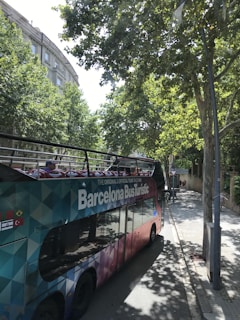 A colorful, double-decker tour bus labeled 'Barcelona Bus Turistic' is parked on a tree-lined street. The bus features an open top with vibrant seating visible. Tall trees cast shadows onto the sidewalk and street, while buildings with classical architecture can be seen in the background.