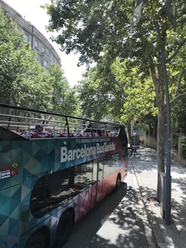 A colorful, double-decker tour bus labeled 'Barcelona Bus Turistic' is parked on a tree-lined street. The bus features an open top with vibrant seating visible. Tall trees cast shadows onto the sidewalk and street, while buildings with classical architecture can be seen in the background.