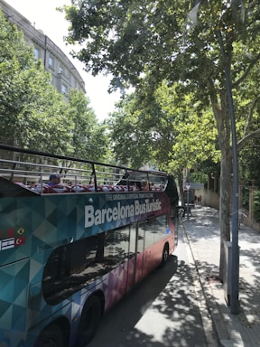 A colorful, double-decker tour bus labeled 'Barcelona Bus Turistic' is parked on a tree-lined street. The bus features an open top with vibrant seating visible. Tall trees cast shadows onto the sidewalk and street, while buildings with classical architecture can be seen in the background.