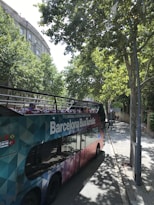 A colorful, double-decker tour bus labeled 'Barcelona Bus Turistic' is parked on a tree-lined street. The bus features an open top with vibrant seating visible. Tall trees cast shadows onto the sidewalk and street, while buildings with classical architecture can be seen in the background.