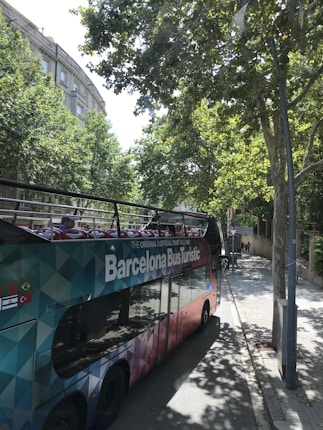 A colorful, double-decker tour bus labeled 'Barcelona Bus Turistic' is parked on a tree-lined street. The bus features an open top with vibrant seating visible. Tall trees cast shadows onto the sidewalk and street, while buildings with classical architecture can be seen in the background.