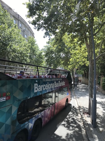 A colorful, double-decker tour bus labeled 'Barcelona Bus Turistic' is parked on a tree-lined street. The bus features an open top with vibrant seating visible. Tall trees cast shadows onto the sidewalk and street, while buildings with classical architecture can be seen in the background.