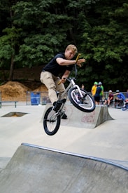 A person is performing a high jump trick on a BMX bike in a skate park. The rider is airborne over a concrete ramp, with one hand off the handlebars showcasing the balance and skill involved. There are several spectators and other riders in the background, with some wearing helmets. The surroundings include trees and a forested area.
