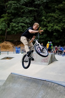 A person is performing a high jump trick on a BMX bike in a skate park. The rider is airborne over a concrete ramp, with one hand off the handlebars showcasing the balance and skill involved. There are several spectators and other riders in the background, with some wearing helmets. The surroundings include trees and a forested area.