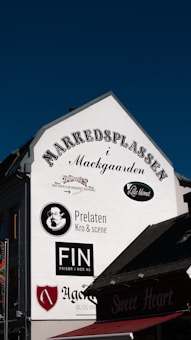 The side of a building features several signs and logos from different businesses. The businesses include Prelaten Kro & Scene, Finn Fris&oslash;r i Nor AS, and several others. The background is a clear blue sky, and part of the roof with the sign 'Sweet Heart' is visible at the bottom.