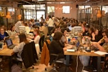 A large group of people are gathered in a spacious, well-lit office or coworking space. They are seated at long wooden tables working on laptops. Some are collaborating in small groups while others are focused on their individual tasks. The atmosphere appears to be busy and productive with a friendly and collaborative vibe. There are decorations like orange balloons and string lights that add to the lively environment.