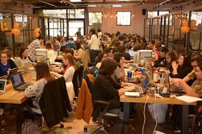 A large group of people are gathered in a spacious, well-lit office or coworking space. They are seated at long wooden tables working on laptops. Some are collaborating in small groups while others are focused on their individual tasks. The atmosphere appears to be busy and productive with a friendly and collaborative vibe. There are decorations like orange balloons and string lights that add to the lively environment.