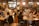 A large group of people are gathered in a spacious, well-lit office or coworking space. They are seated at long wooden tables working on laptops. Some are collaborating in small groups while others are focused on their individual tasks. The atmosphere appears to be busy and productive with a friendly and collaborative vibe. There are decorations like orange balloons and string lights that add to the lively environment.