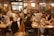 A large group of people are gathered in a spacious, well-lit office or coworking space. They are seated at long wooden tables working on laptops. Some are collaborating in small groups while others are focused on their individual tasks. The atmosphere appears to be busy and productive with a friendly and collaborative vibe. There are decorations like orange balloons and string lights that add to the lively environment.