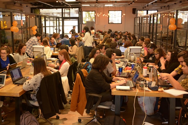 A large group of people are gathered in a spacious, well-lit office or coworking space. They are seated at long wooden tables working on laptops. Some are collaborating in small groups while others are focused on their individual tasks. The atmosphere appears to be busy and productive with a friendly and collaborative vibe. There are decorations like orange balloons and string lights that add to the lively environment.