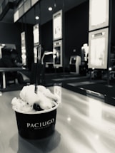 A cup of gelato labeled Paciugo sits on a table with a spoon in it. The background includes seating and wall displays, with a person in the distance.
