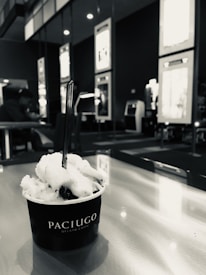 A cup of gelato labeled Paciugo sits on a table with a spoon in it. The background includes seating and wall displays, with a person in the distance.