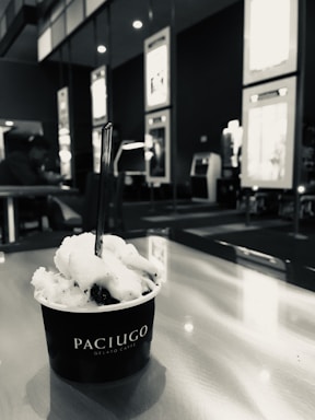 A cup of gelato labeled Paciugo sits on a table with a spoon in it. The background includes seating and wall displays, with a person in the distance.