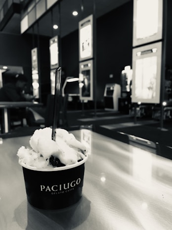 A cup of gelato labeled Paciugo sits on a table with a spoon in it. The background includes seating and wall displays, with a person in the distance.