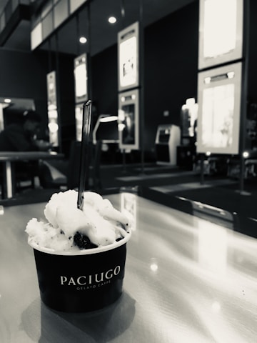 A cup of gelato labeled Paciugo sits on a table with a spoon in it. The background includes seating and wall displays, with a person in the distance.