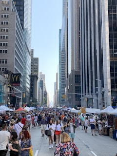 A bustling city street, lined with tall skyscrapers and filled with numerous people walking and browsing various market stalls. Flags and colorful tents are visible, suggesting a lively street fair or market event. The buildings on either side display a mix of modern glass facades and more traditional architecture.