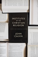 A black book with the title 'Institutes of the Christian Religion' by John Calvin is prominently placed on top of several open books with visible text. The scene suggests a scholarly or religious study setting.