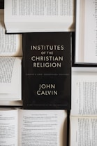A black book with the title 'Institutes of the Christian Religion' by John Calvin is prominently placed on top of several open books with visible text. The scene suggests a scholarly or religious study setting.