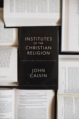 A black book with the title 'Institutes of the Christian Religion' by John Calvin is prominently placed on top of several open books with visible text. The scene suggests a scholarly or religious study setting.