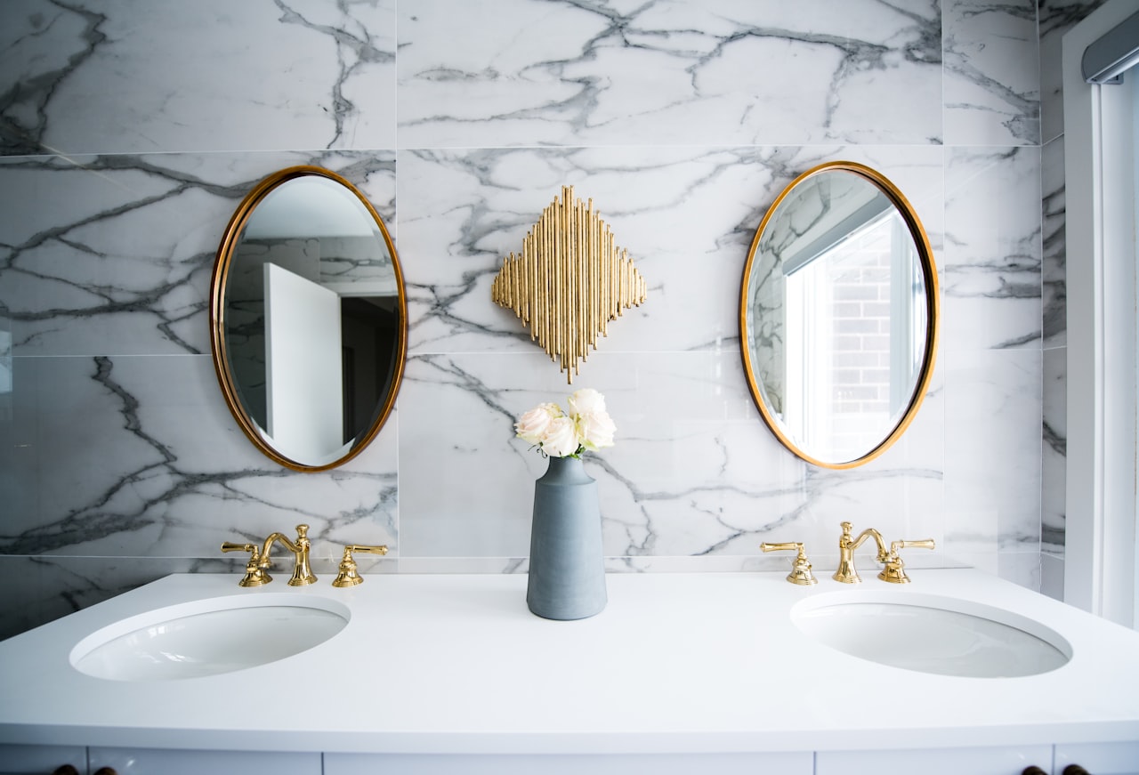 Retro, 1920s-style of bathroom with a dual vanity. Marble tiled walls and brass mirrors and faucets. A brass art deco wall sculpture hangs between the two mirrors.