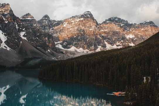 A serene mountain landscape with towering, snow-capped peaks reflecting on a calm, turquoise lake. Dense evergreen forests surround the lake, adding depth to the scene. Several brightly colored canoes or kayaks are lined up along the shoreline.
