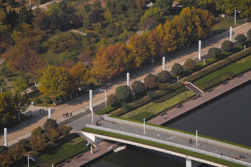 An aerial view of a park featuring a long pathway lined with tall white light poles and trees with autumn foliage. There are landscaped grassy areas alongside the path and a canal or river with a bridge crossing over it. Several people are walking along the path, and the scene is peaceful and well-maintained.