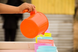 A hand holds an orange jug, pouring water into a row of colorful plastic cups arranged neatly. The cups are in bright shades of pink, blue, and yellow, and are lined up on a surface with a blurred background featuring a yellow and gray structure.