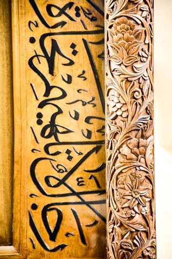 A detailed wooden panel features ornate floral carvings on the right with intricate patterns, while elegant calligraphic script in black ink adorns the left side of the panel, contrasting beautifully with the wood's natural tones.