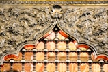 An intricately designed metallic structure featuring elaborate floral patterns and ornate details. The design includes a repetitive pattern of small, spherical connectors forming a grid, with a prominent arch shape adorned with leaves and floral motifs. The metal is polished, giving it a reflective sheen, and it are accentuated by a warm, orange glow coming from the background.