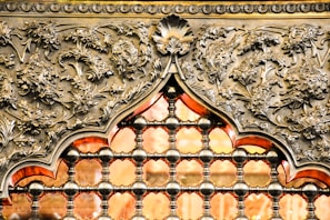 An intricately designed metallic structure featuring elaborate floral patterns and ornate details. The design includes a repetitive pattern of small, spherical connectors forming a grid, with a prominent arch shape adorned with leaves and floral motifs. The metal is polished, giving it a reflective sheen, and it are accentuated by a warm, orange glow coming from the background.
