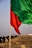 A large green flag in the foreground and a red flag mounted on a wooden pole in the midground, both waving in the wind. The background shows a blurred crowd of people walking on a dirt path with a clear sky above.