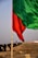 A large green flag in the foreground and a red flag mounted on a wooden pole in the midground, both waving in the wind. The background shows a blurred crowd of people walking on a dirt path with a clear sky above.