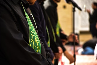 Several people are standing in a solemn formation, wearing black garments with green sashes that have golden inscriptions. They appear to be engaged in a ceremonial or religious gathering, and a microphone is visible, indicating possible speeches or prayers.