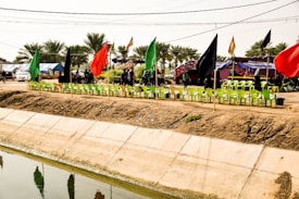 A line of green chairs is arranged alongside a dirt path with vibrant red, green, and black flags. A tent with banners and people is visible in the background. Palm trees are in the distance, and there's a canal in the foreground.