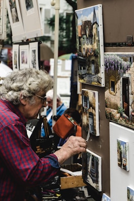 An elderly man with curly gray hair and a red plaid shirt focuses on painting a small artwork on a display board. The surrounding area is filled with other framed paintings featuring detailed building facades and scenic views. The scene suggests an outdoor art market or fair.