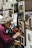 An elderly man with curly gray hair and a red plaid shirt focuses on painting a small artwork on a display board. The surrounding area is filled with other framed paintings featuring detailed building facades and scenic views. The scene suggests an outdoor art market or fair.