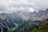 A paraglider is soaring over a breathtaking mountainous landscape. The scene is dominated by rugged peaks and steep cliffs, with patches of green vegetation contrasting against the gray rock. The sky is overcast with heavy clouds, casting a dramatic light over the scenery.