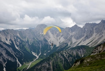 A paraglider is soaring over a breathtaking mountainous landscape. The scene is dominated by rugged peaks and steep cliffs, with patches of green vegetation contrasting against the gray rock. The sky is overcast with heavy clouds, casting a dramatic light over the scenery.