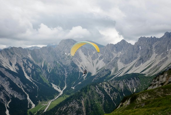 A paraglider is soaring over a breathtaking mountainous landscape. The scene is dominated by rugged peaks and steep cliffs, with patches of green vegetation contrasting against the gray rock. The sky is overcast with heavy clouds, casting a dramatic light over the scenery.