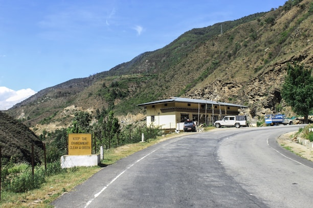 A winding road leads past a small building with several parked vehicles, including a white truck and a blue truck. The landscape is mountainous, with green hills and rocky terrain surrounding the area. A sign on the left reads 'KEEP THE ENVIRONMENT CLEAN & GREEN', emphasizing environmental consciousness.