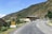A winding road leads past a small building with several parked vehicles, including a white truck and a blue truck. The landscape is mountainous, with green hills and rocky terrain surrounding the area. A sign on the left reads 'KEEP THE ENVIRONMENT CLEAN & GREEN', emphasizing environmental consciousness.