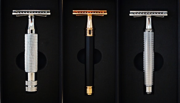 Three safety razors are neatly displayed in a black case. The razors have metallic handles with two silver and one gold in color. The middle razor has a sleek black handle with gold accents, while the side razors have textured silver handles.
