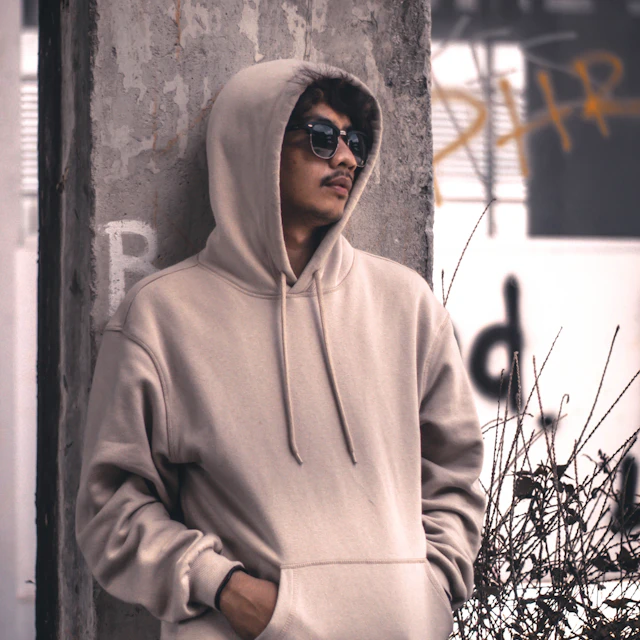 A man in a grey hoodie leaning against a wall in daylight