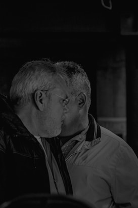 Two men are facing each other closely, partially obscured, in a dark, moody setting. They are wearing jackets and appear to be engaged in a private conversation.