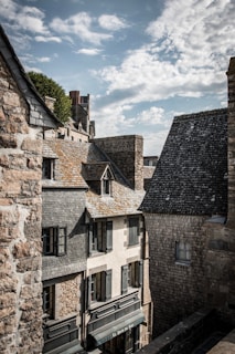 Historic stone buildings with sloped roofs and wooden shutters are closely arranged under a partly cloudy sky. The textures of the stone and shingles convey a sense of age and heritage.