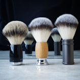 A set of three shaving brushes with different handles arranged on a reflective surface, their soft bristles splayed outwards. The handle materials range from black and glossy, to a wooden finish and a sleek metallic look. The background is dark, contrasting with the lighter bristles of the brushes.
