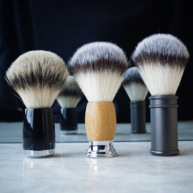 A set of three shaving brushes with different handles arranged on a reflective surface, their soft bristles splayed outwards. The handle materials range from black and glossy, to a wooden finish and a sleek metallic look. The background is dark, contrasting with the lighter bristles of the brushes.