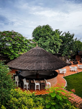 A thatched roof structure acts as a circular outdoor bar surrounded by high chairs. The setting includes lush greenery with trees and bushes. Brick pathways weave through an outdoor seating area with round tables and chairs.